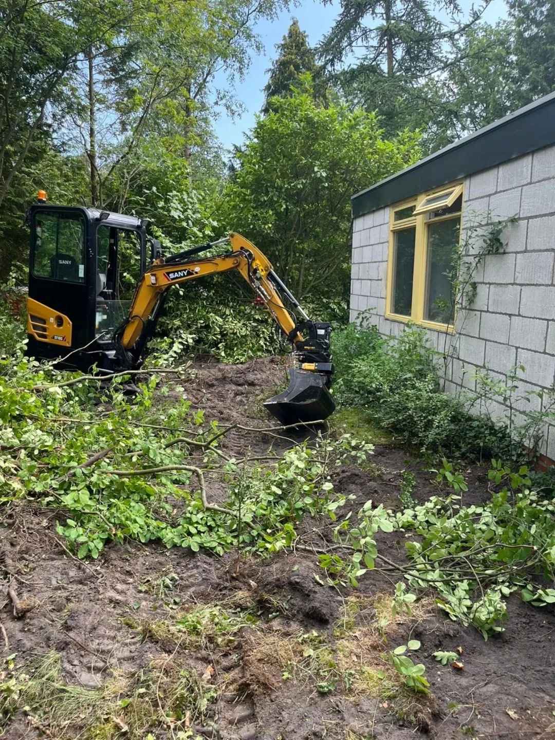 Kleine graafmachine ruimt struiken en bosschage op naast een gebouw